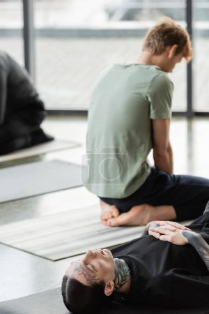 Tattooed man lying on mat near blurred people in yoga class 