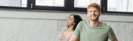 Positive redhead man looking at camera near middle eastern woman in yoga studio, banner 