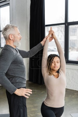 Entraîneur d'âge moyen debout près d'une femme afro-américaine faisant Crescent Lunge asana en cours de yoga 