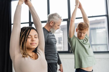 Téléchargez les photos : Jeune femme afro-américaine méditant à Crescent Lunge asana près d'un groupe flou dans un studio de yoga - en image libre de droit