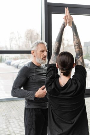 Mature coach talking to tattooed man doing Crescent Lunge asana in yoga studio 