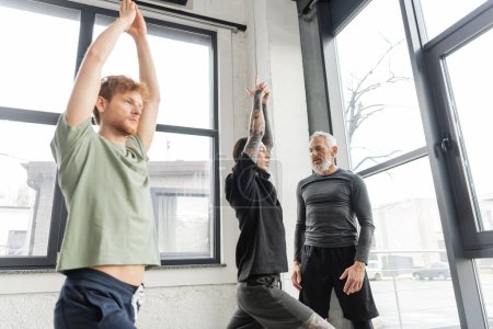 Entraîneur d'âge moyen parlant à des jeunes hommes faisant Crescent Lunge asana dans un studio de yoga 