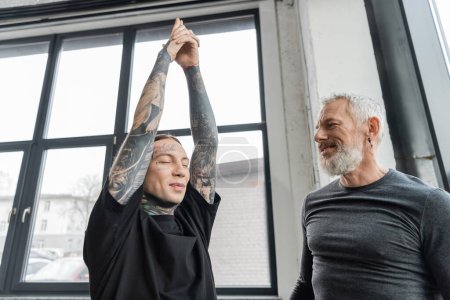 Lächelnder Trainer mittleren Alters schaut tätowierten Mann bei Crescent Lunge Asana im Yoga-Kurs an 