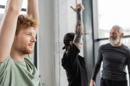 Smiling redhead man standing in Crescent Lunge asana near blurred group in yoga studio 