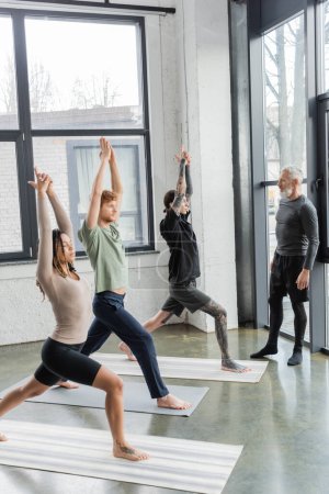 Personas interraciales de pie en Crescent Lunge asana cerca de entrenador maduro en clase de yoga 