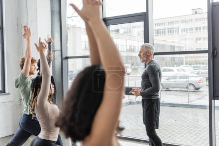 Téléchargez les photos : Vue latérale du coach mature parlant à des personnes multiethniques debout dans la pose de Crescent Lunge dans un studio de yoga - en image libre de droit