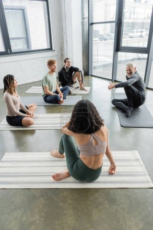 Smiling middle aged coach talking to interracial group of people on mats in yoga class 
