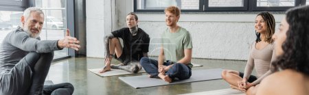 Smiling coach pointing at middle eastern woman near multiethnic group in yoga class, banner 