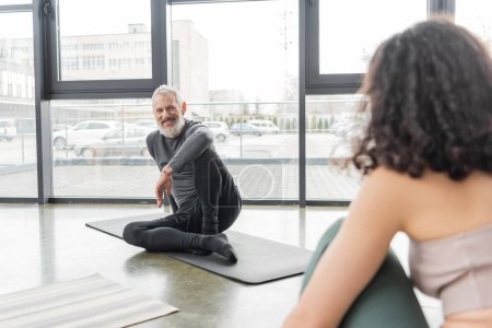 Cheerful mature coach looking at blurred woman while sitting on mat in yoga studio 