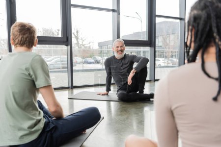 Positive coach looking at blurred multiethnic people in yoga studio 