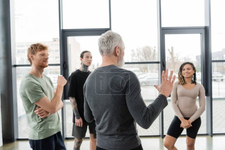 Mature coach gesturing near young interracial people in yoga class 