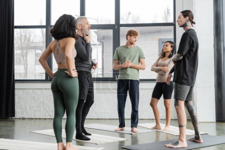 Personas multiétnicas hablando mientras están de pie sobre alfombras en el estudio de yoga 