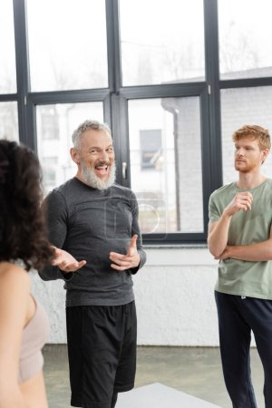 Positive mature coach pointing at blurred woman in yoga studio 