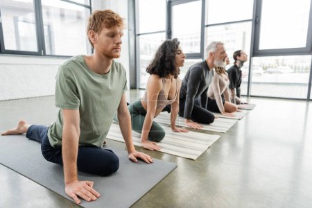 Rotschopf praktiziert Half Pigeon Asana in der Nähe multiethnischer Menschen im Yoga-Kurs 