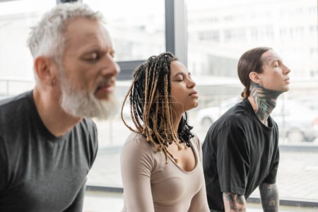 Téléchargez les photos : Jeune femme afro-américaine méditant les yeux fermés près des hommes flous dans un studio de yoga - en image libre de droit