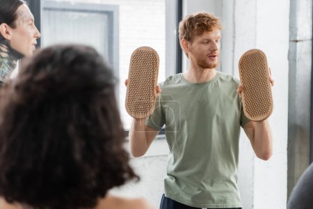 Young redhead man looking at sadhu board and talking to people in yoga class 