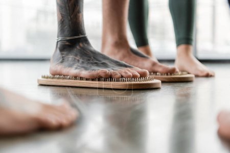 częściowy widok człowieka z tatuażem na nogach stojącego na desce do paznokci w pobliżu osób w studio jogi