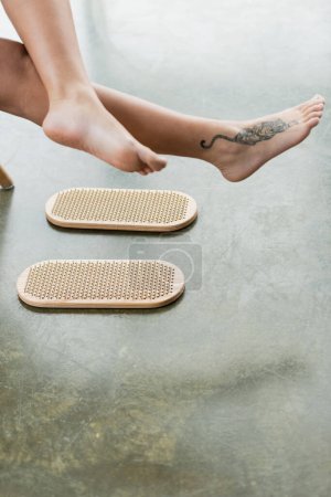 Teilaufnahme einer tätowierten Frau mit nackten Füßen in der Nähe von Sadhu Board im Yoga-Studio 