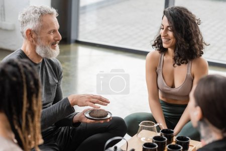 hombre tatuado con barba gris sosteniendo té puer comprimido cerca de alegre mujer de Oriente Medio en estudio de yoga 
