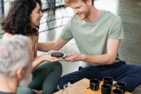 happy middle eastern woman giving compressed puer tea to smiling redhead man in yoga studio 