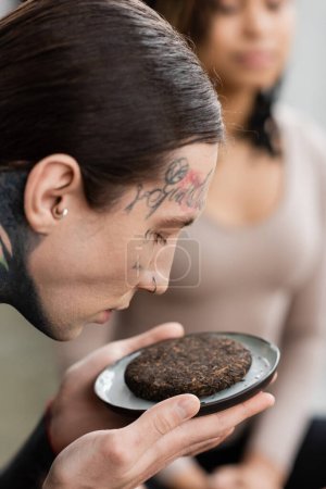 Foto de Hombre joven y tatuado oliendo té puer fermentado cerca de una mujer afroamericana sobre un fondo borroso - Imagen libre de derechos