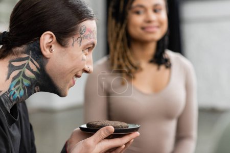happy and tattooed man holding fermented puer tea near african american woman on blurred background 