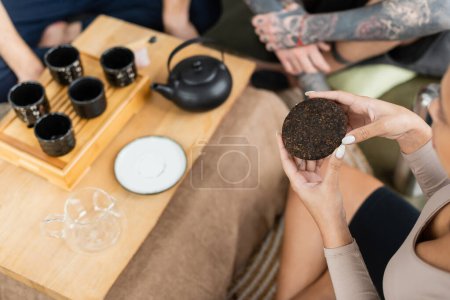vista aérea de la mujer afroamericana sosteniendo té puro comprimido cerca de la gente y la olla de té tradicional sobre fondo borroso 