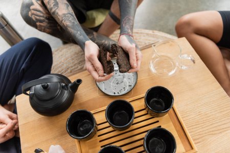 Draufsicht des tätowierten Mannes, der komprimierten Puer-Tee in der Nähe traditioneller chinesischer Teekanne und Tassen bricht 
