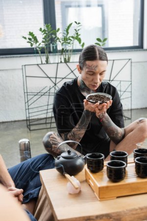 young man with tattoos smelling fermented puer tea near burning Palo Santo stick and Chinese teapot with cups 