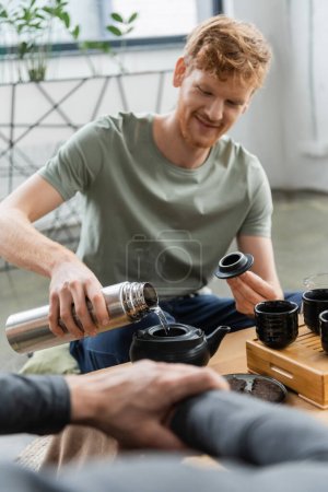 happy redhead man holding thermos while pouring hot water and brewing puer tea in Chinese tea pot 