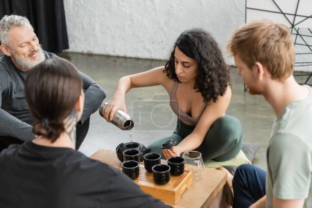 Photo for Curly middle eastern woman pouring hot water from thermos and brewing puer tea near men in yoga studio - Royalty Free Image