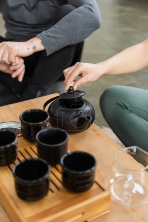 abgeschnittene Ansicht einer Frau, die Tee in einer chinesischen Teekanne in der Nähe tätowierter Mann mittleren Alters braut 