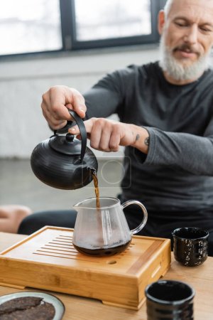 tattooed middle aged man pouring freshly brewed puer tea into glass jug 