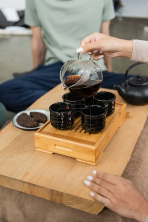 partial view of woman pouring brewed puer from glass jug into traditional tea cups 