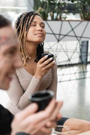 mujer afroamericana complacida con rastas sosteniendo la taza con té puro cerca del hombre tatuado en primer plano borroso