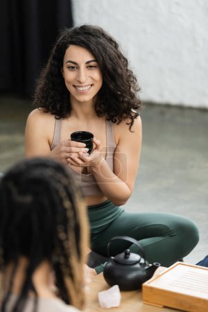happy middle eastern woman with curly hair holding Chinese cup during tea ceremony in yoga studio 