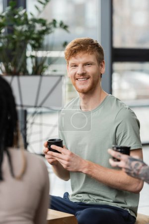 happy man with red hair holding Japanese cup with puer tea near people in yoga studio 