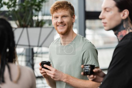 Photo for Cheerful man with red hair holding japanese cup with puer tea near people in yoga studio - Royalty Free Image