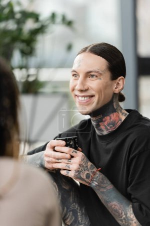 hombre feliz y tatuado sonriendo mientras sostiene la taza japonesa con té cerca de la mujer en primer plano borroso 