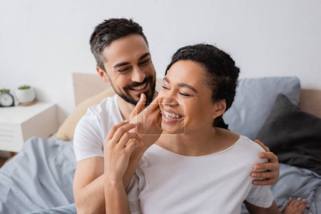 smiling bearded man touching face of overjoyed african american woman in bedroom at home