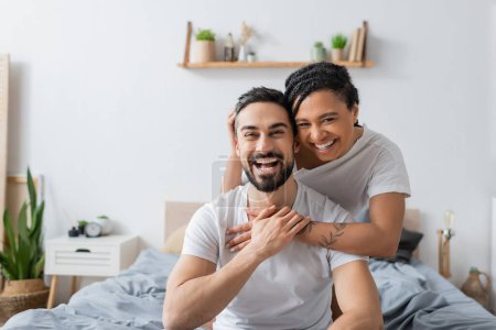 Überglückliches multiethnisches Paar in weißen T-Shirts lacht im heimischen Schlafzimmer in die Kamera