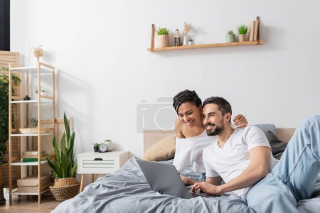 Unbekümmerte interrassische Paare lächeln in der Nähe von Laptop, während Film im modernen Schlafzimmer zu Hause