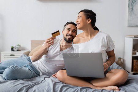 Téléchargez les photos : Heureux barbu homme tenant carte de crédit près de rire afro-américaine petite amie assis avec ordinateur portable sur le lit à la maison - en image libre de droit