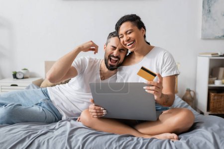 Foto de Pareja interracial llena de alegría riendo con los ojos cerrados cerca de la computadora portátil y la tarjeta de crédito en la cama en casa - Imagen libre de derechos