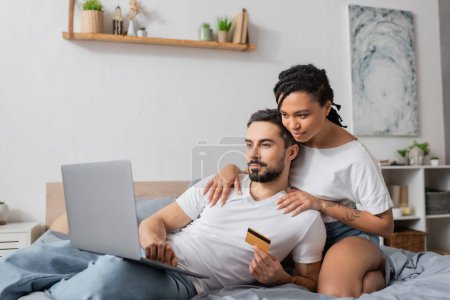 joven afroamericano mujer abrazando hombros de barbudo hombre con portátil y tarjeta de crédito en la cama en casa