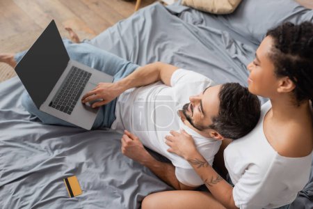vue du haut de sourire homme barbu en utilisant un ordinateur portable près de petite amie afro-américaine et carte de crédit sur le lit à la maison