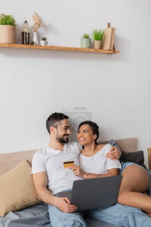 Téléchargez les photos : Heureux couple interracial avec ordinateur portable et carte de crédit se regardant dans la chambre à coucher à la maison - en image libre de droit