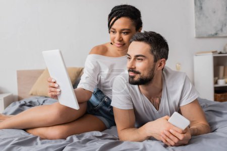 Lächelnde Afroamerikanerin zeigt ihrem bärtigen Freund, der mit Smartphone auf dem Bett liegt, ein digitales Tablet