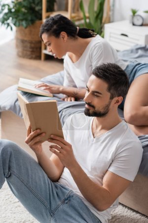 hombre barbudo positivo y mujer afroamericana joven leyendo libros en el dormitorio en casa