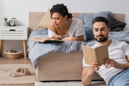 young african american woman and bearded man reading books while resting in bedroom at home